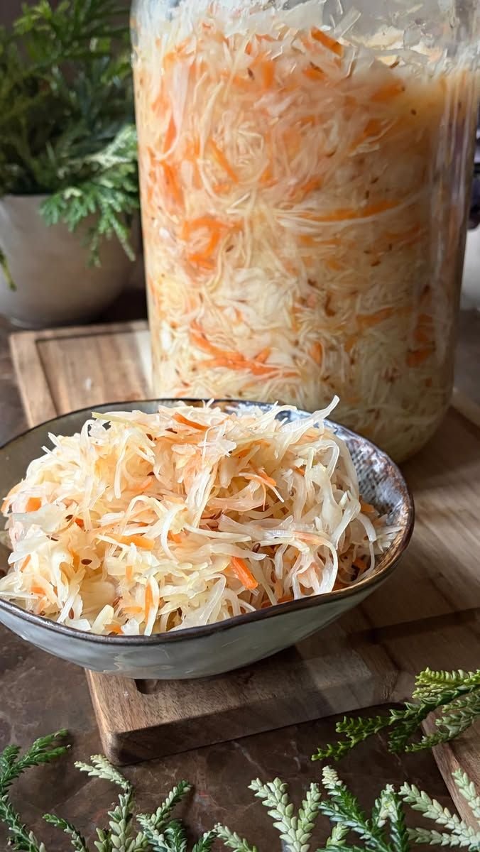 A bowl of sauerkraut with carrot in front of a jar of fermenting cabbage